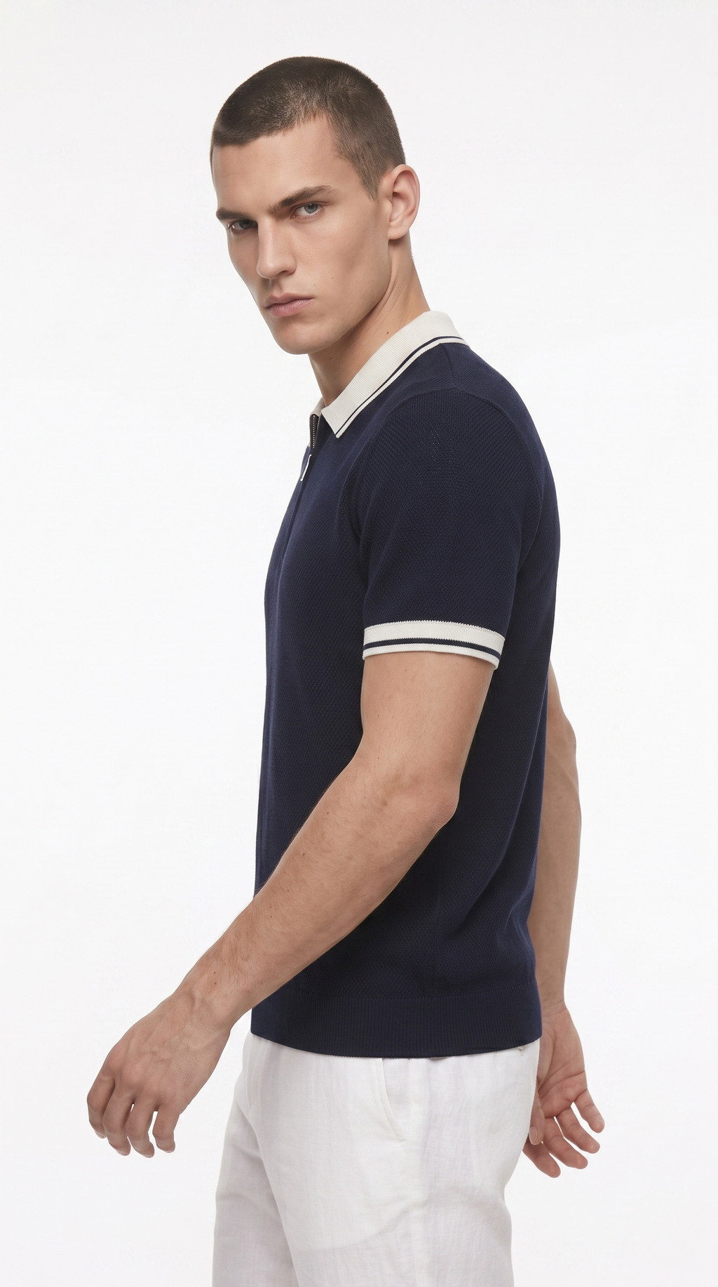 A man wearing a navy blue short-sleeve polo shirt with white stripes on the collar and sleeve cuffs, paired with white trousers, photographed against a plain white studio background, from Meleche Fashion USA or Meléche (short as (Mé))