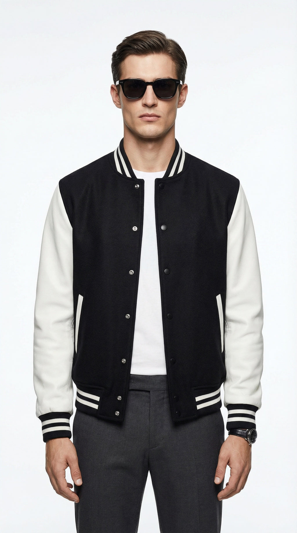 Man wearing a black and white varsity jacket, white t-shirt, and dark jeans, holding a black leather jacket, standing against a gray wall.