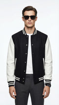 Man wearing a black and white varsity jacket, white t-shirt, and dark jeans, holding a black leather jacket, standing against a gray wall.