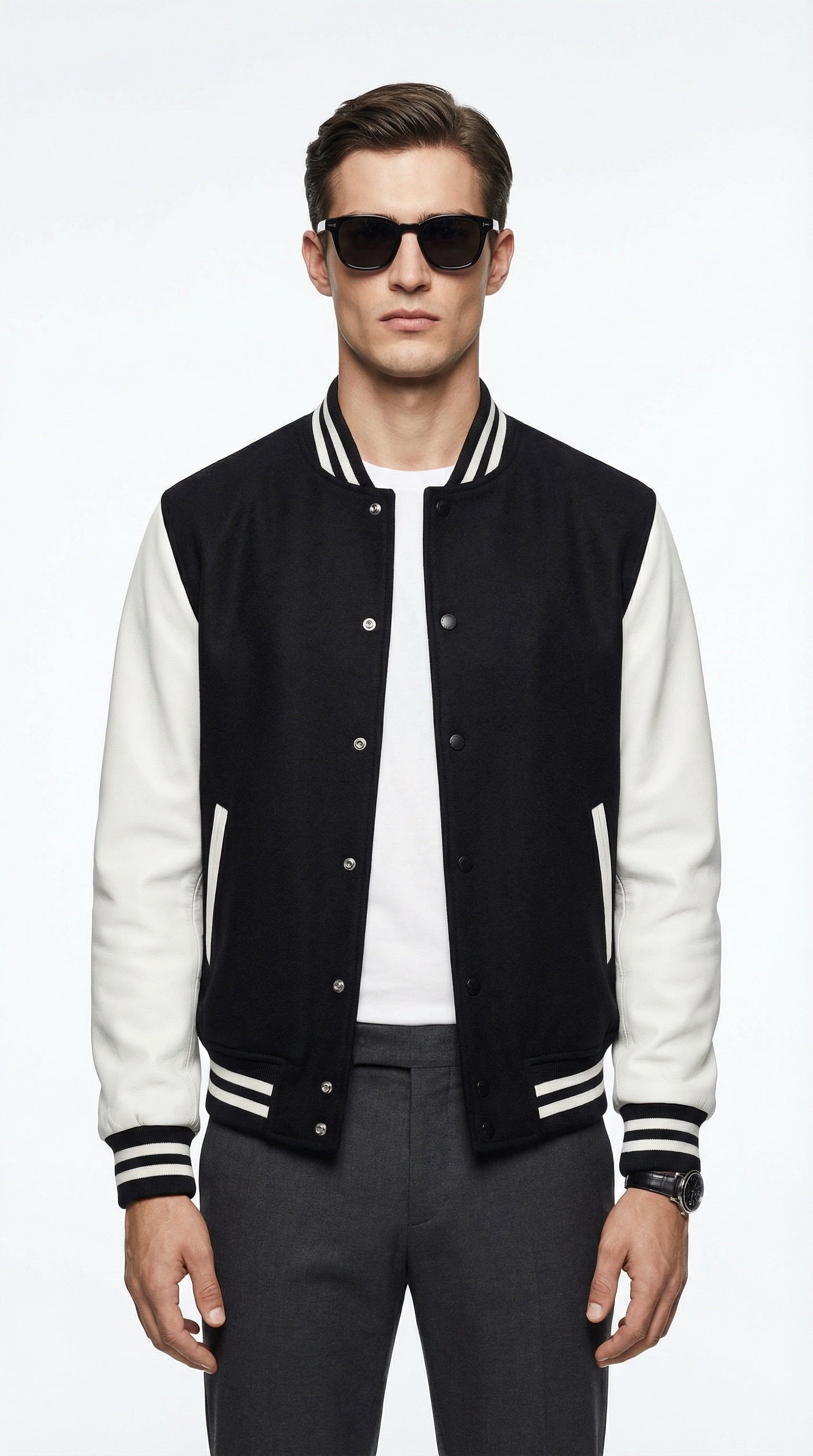 Man wearing a black and white varsity jacket, white t-shirt, and dark jeans, holding a black leather jacket, standing against a gray wall.