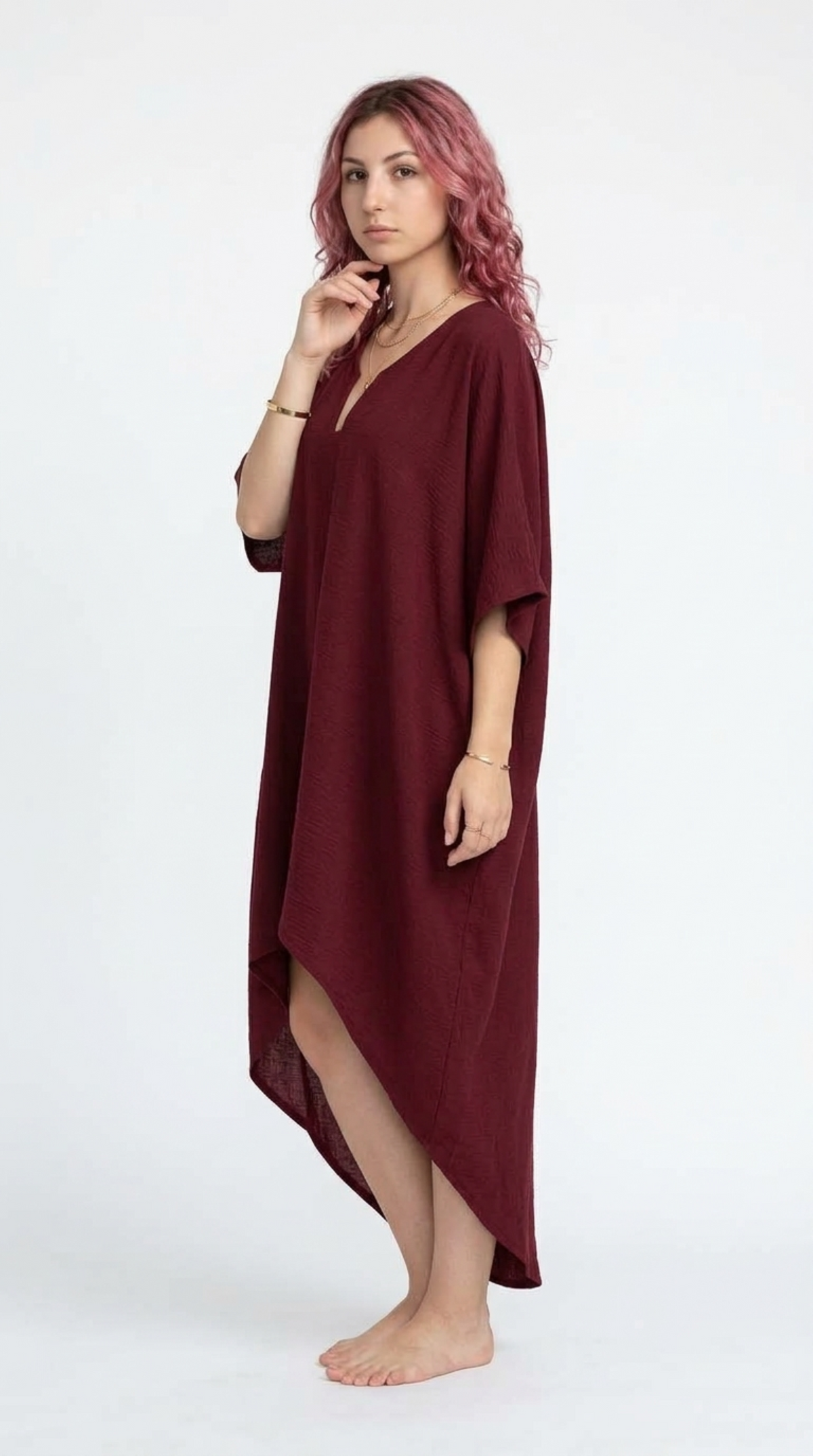 A woman with pink hair stands barefoot against a plain white studio background, wearing a loose, draped maxi cover-up dress in deep burgundy with a V-neck and elbow-length sleeves, from Meleche Fashion or Meléche (short as (Mé)) and shoot taken by Meléche studio photography NYC-USA