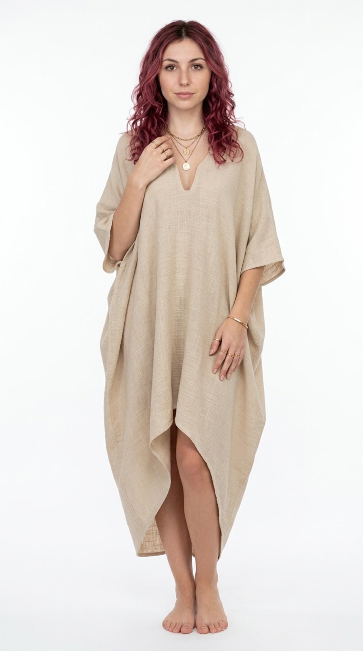 A woman with wavy reddish hair stands barefoot against a white studio background, wearing a loose, draped kaftan-style cover-up dress in a light warm beige linen-like fabric, from Meleche Fashion or Meléche (short as (Mé)) and shoot taken by Meléche studio photography NYC-USA
