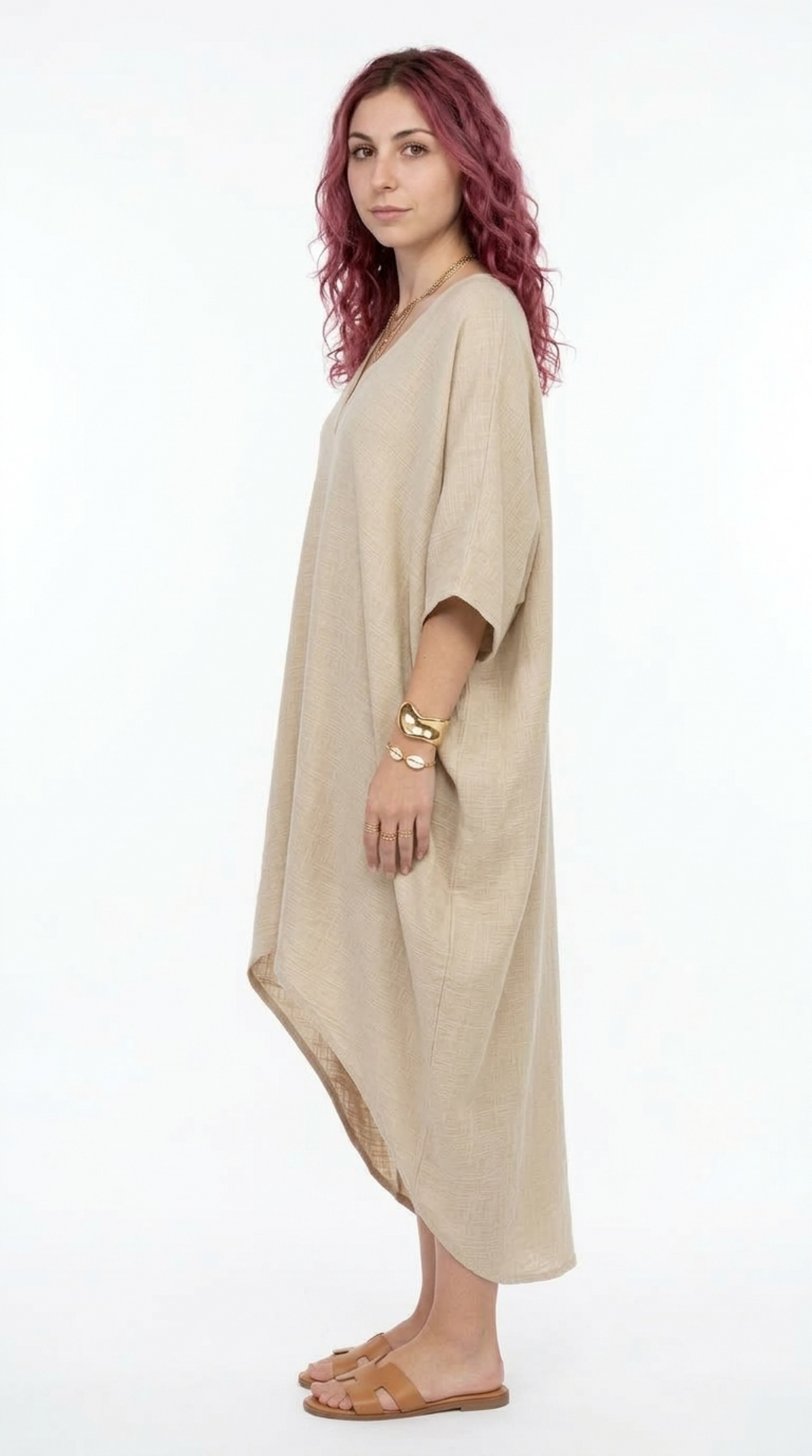 A woman with wavy magenta hair stands in profile wearing a loose, draped kaftan-style dress in a light beige linen-like fabric. She is wearing tan flat sandals and gold bracelets against a plain white studio background, from Meleche Fashion or Meléche (short as (Mé)) and shoot taken by Meléche studio photography NYC-USA