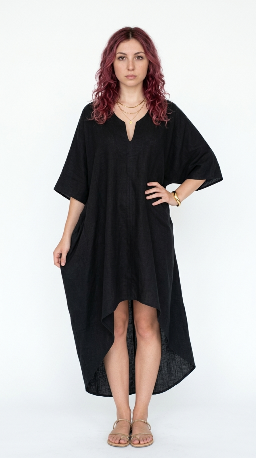 A woman with wavy red hair wearing a loose, black high-low kaftan dress with wide three-quarter sleeves and tan flat sandals, standing against a plain white studio background, from Meleche Fashion or Meléche (short as (Mé)) and shoot taken by Meléche studio photography NYC-USA