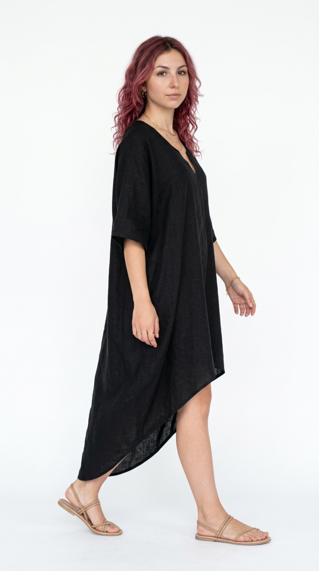 A woman modeling a loose, black, short-sleeve high-low dress or kaftan with a V-neckline, paired with beige flat sandals, against a plain white studio background, from Meleche Fashion or Meléche (short as (Mé)) and shoot taken by Meléche studio photography NYC-USA