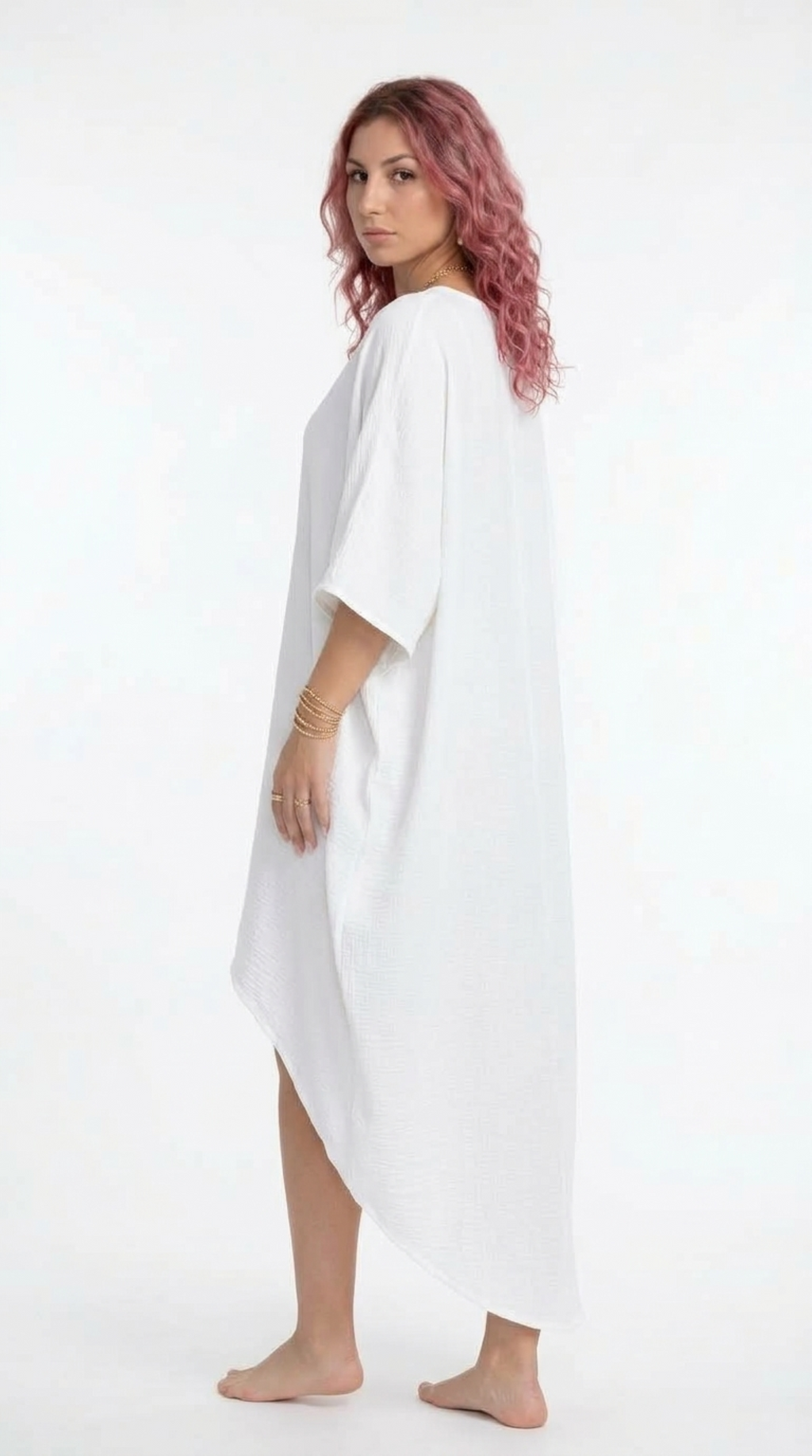 A woman with pink hair stands barefoot in a white, loose-fitting kaftan-style dress or cover-up against a plain white studio background, from Meleche Fashion or Meléche (short as (Mé)) and shoot taken by Meléche studio photography NYC-USA
