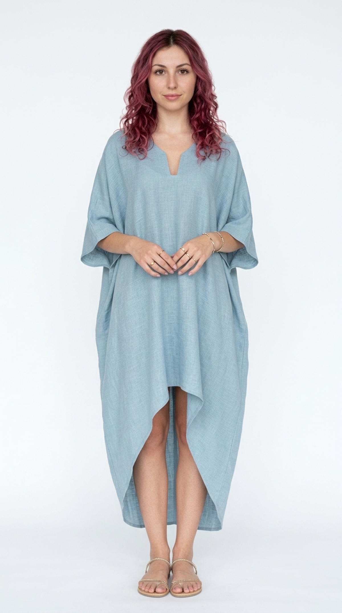 A woman with pinkish-red curly hair wearing a light blue linen kaftan dress with a split high-low hem and wide three-quarter sleeves, standing against a white studio background, from Meleche Fashion or Meléche (short as (Mé)) and shoot taken by Meléche studio photography NYC-USA