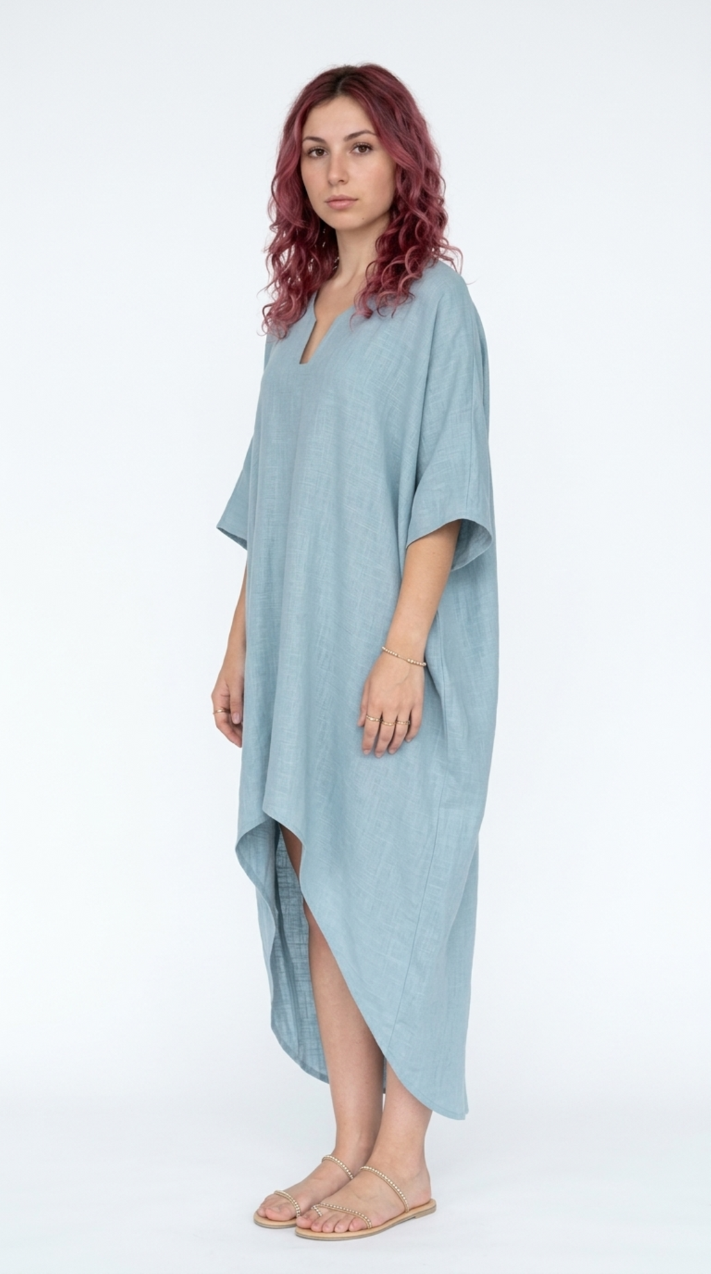 A woman with wavy reddish hair models a light blue linen caftan dress with a split high-low hem and wide short sleeves against a plain white studio background, from Meleche Fashion or Meléche (short as (Mé)) and shoot taken by Meléche studio photography NYC-USA