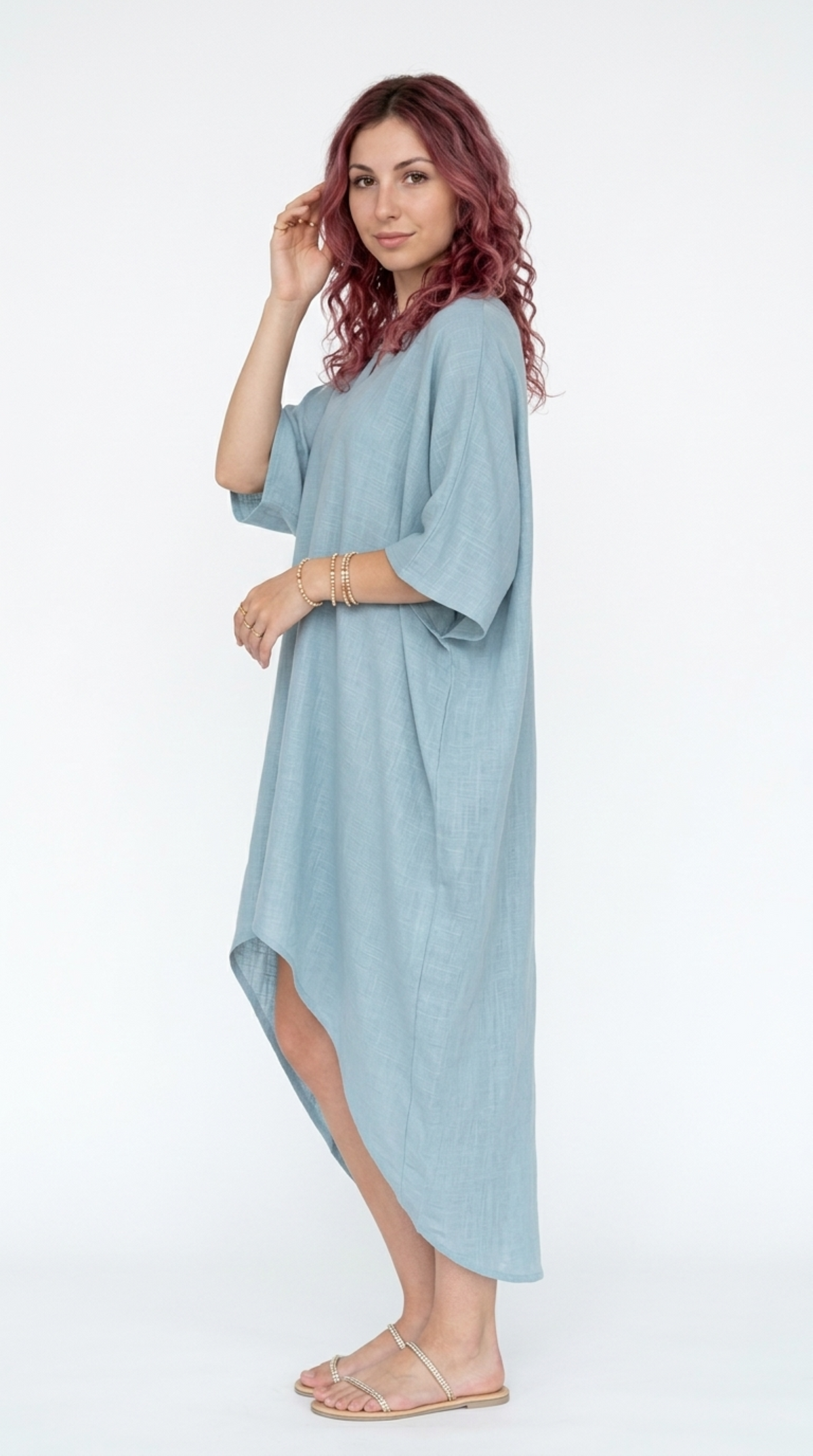 A woman modeling a light blue linen kaftan dress with three-quarter sleeves and an asymmetrical high-low hem, standing against a plain white studio background, from Meleche Fashion or Meléche (short as (Mé)) and shoot taken by Meléche studio photography NYC-USA