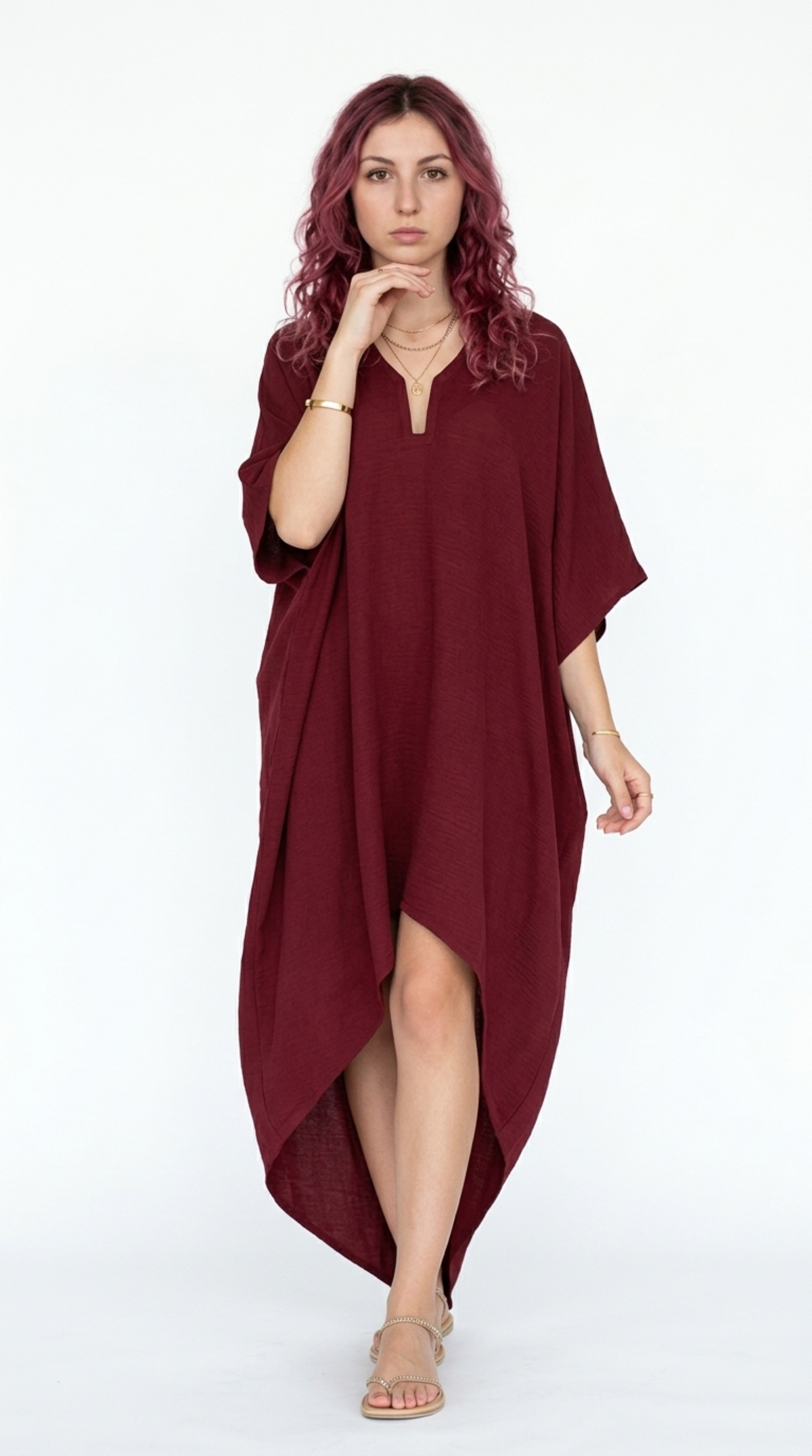 A woman with wavy pink hair wearing a loose, high-low burgundy kaftan dress and beige strappy sandals, posing against a solid white studio background, from Meleche Fashion or Meléche (short as (Mé)) and shoot taken by Meléche studio photography NYC-USA