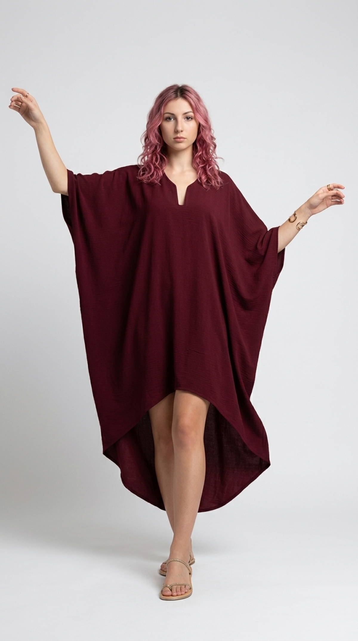 A woman with pink hair models a loose-fitting, high-low length burgundy poncho dress with wide short sleeves and a split neckline, set against a plain light gray studio background, from Meleche Fashion or Meléche (short as (Mé)) and shoot taken by Meléche studio photography NYC-USA