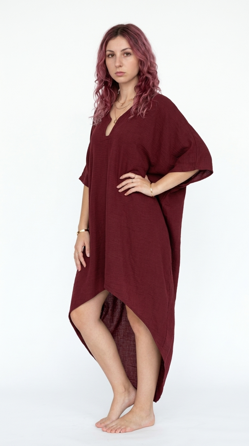 A woman with pinkish-purple hair stands barefoot against a white studio background wearing a loose, high-low length burgundy dress with wide short sleeves, from Meleche Fashion or Meléche (short as (Mé)) and shoot taken by Meléche studio photography NYC-USA