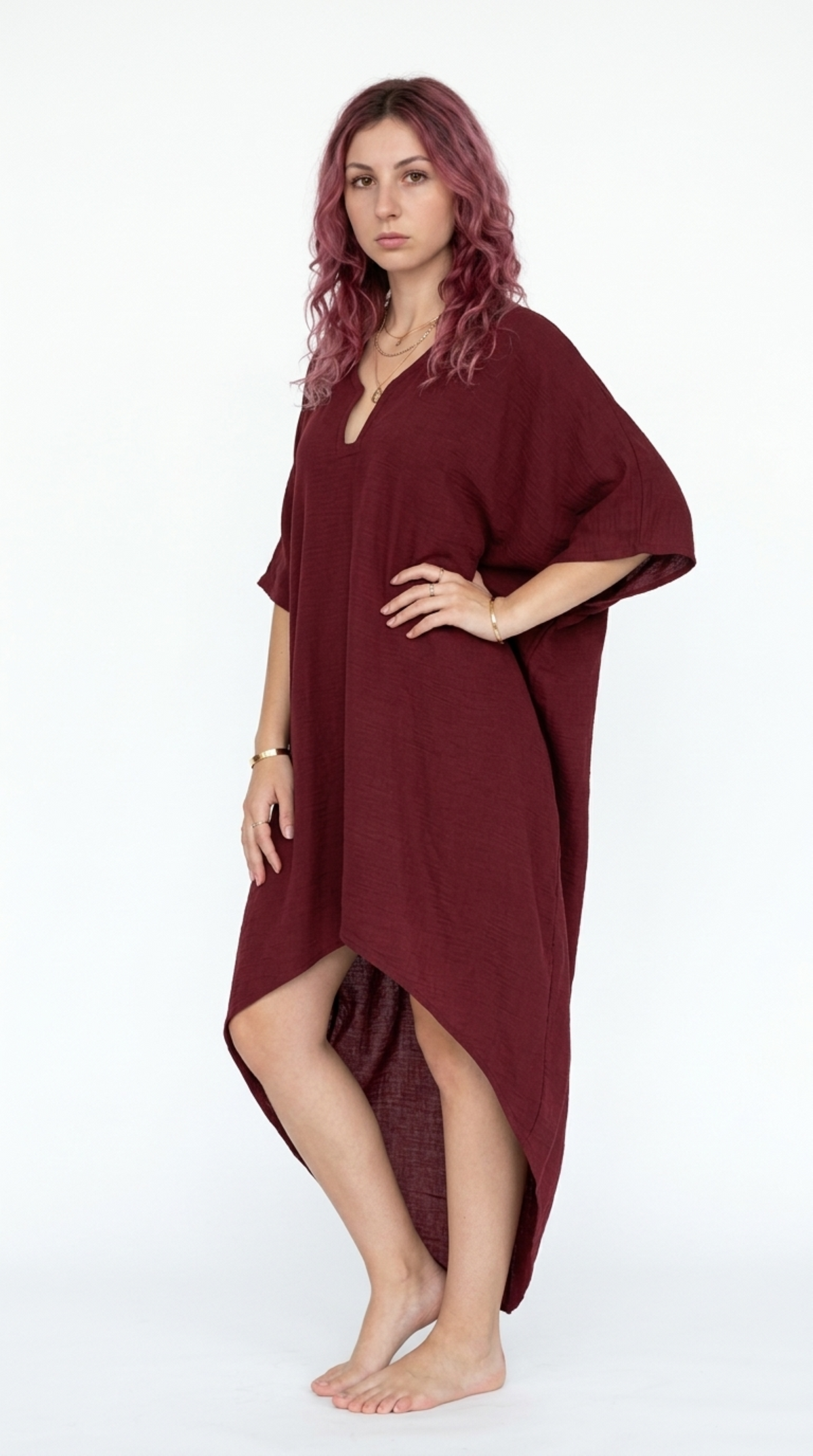 A woman with pinkish-purple hair stands barefoot against a white studio background wearing a loose, high-low length burgundy dress with wide short sleeves, from Meleche Fashion or Meléche (short as (Mé)) and shoot taken by Meléche studio photography NYC-USA