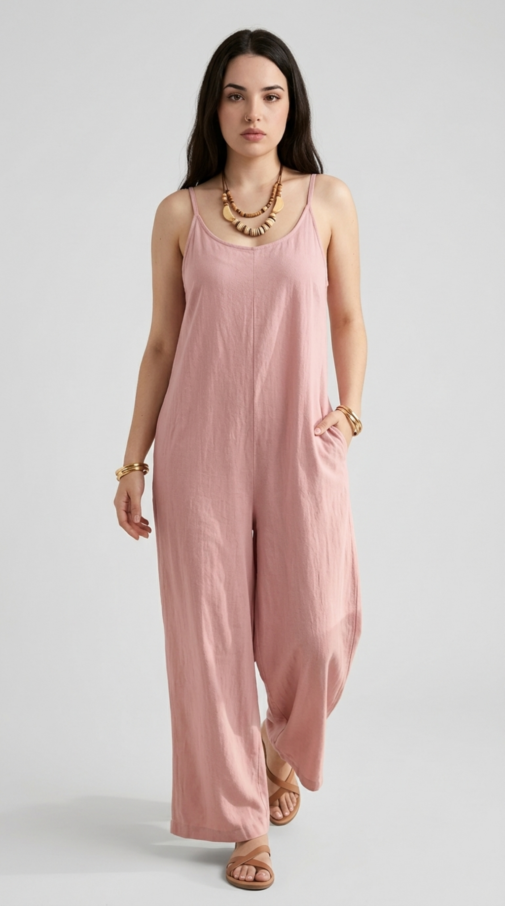 A woman modeling a dusty pink sleeveless jumpsuit with wide legs, styled with a chunky gold necklace and bracelets against a light gray studio background, from Meleche Fashion or Meléche (short as (Mé)) and shoot taken by Meléche studio photography NYC-USA