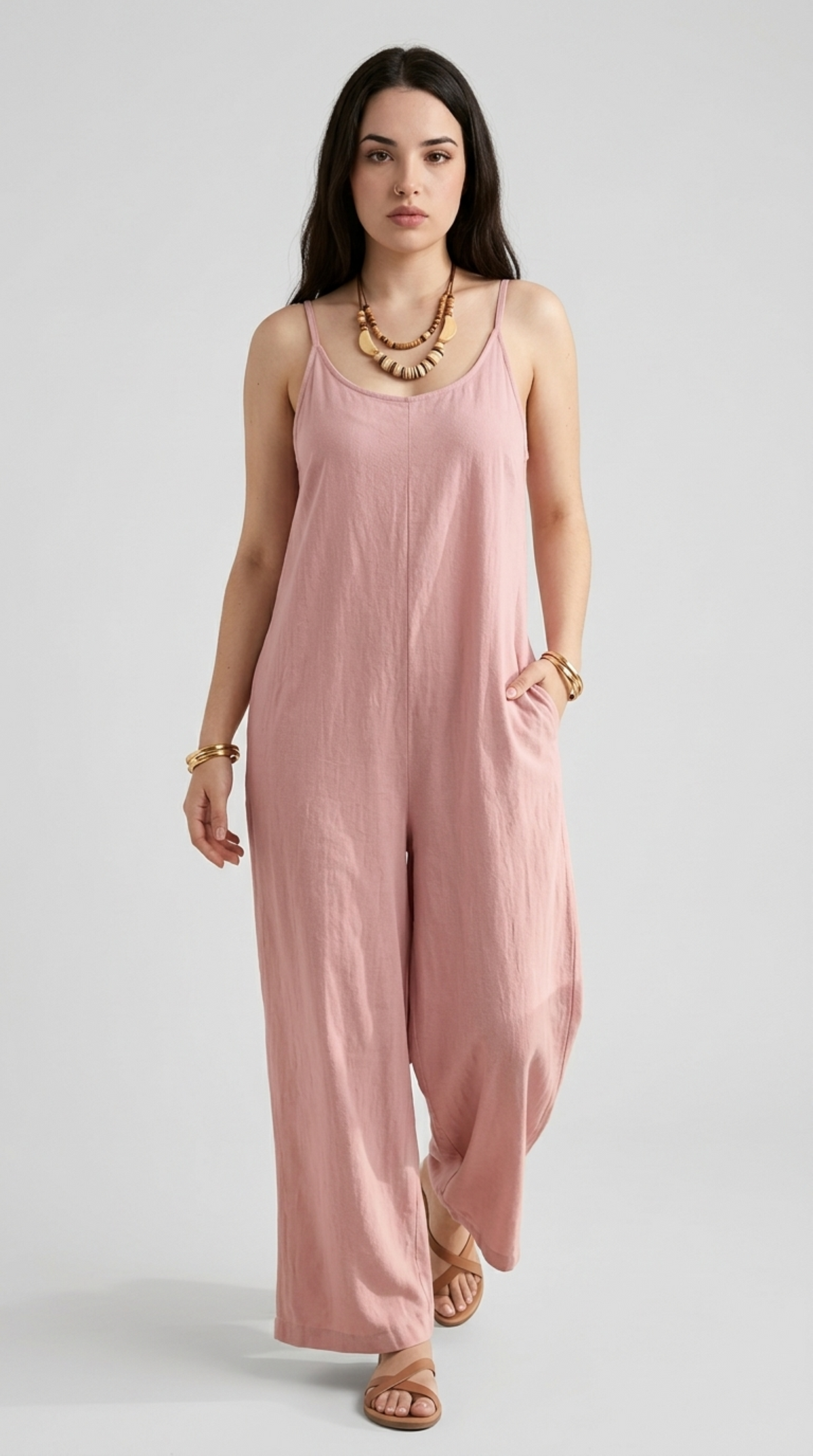 A woman modeling a dusty pink sleeveless jumpsuit with wide legs, styled with a chunky gold necklace and bracelets against a light gray studio background, from Meleche Fashion or Meléche (short as (Mé)) and shoot taken by Meléche studio photography NYC-USA