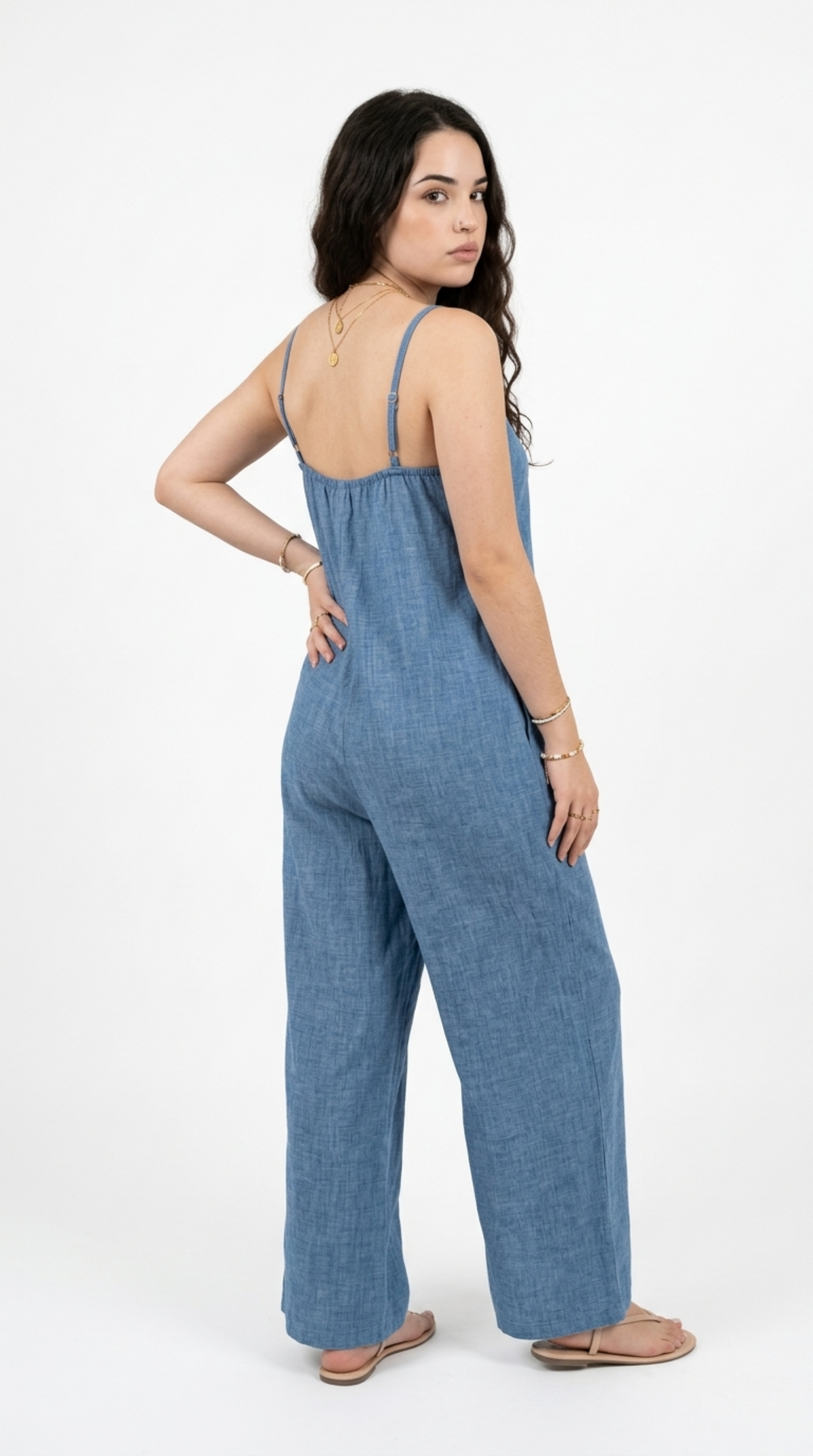 A woman modeling a blue denim wide-leg jumpsuit, shown from a three-quarters back view against a plain white studio background, from Meleche Fashion or Meléche (short as (Mé)) and shoot taken by Meléche studio photography NYC-USA