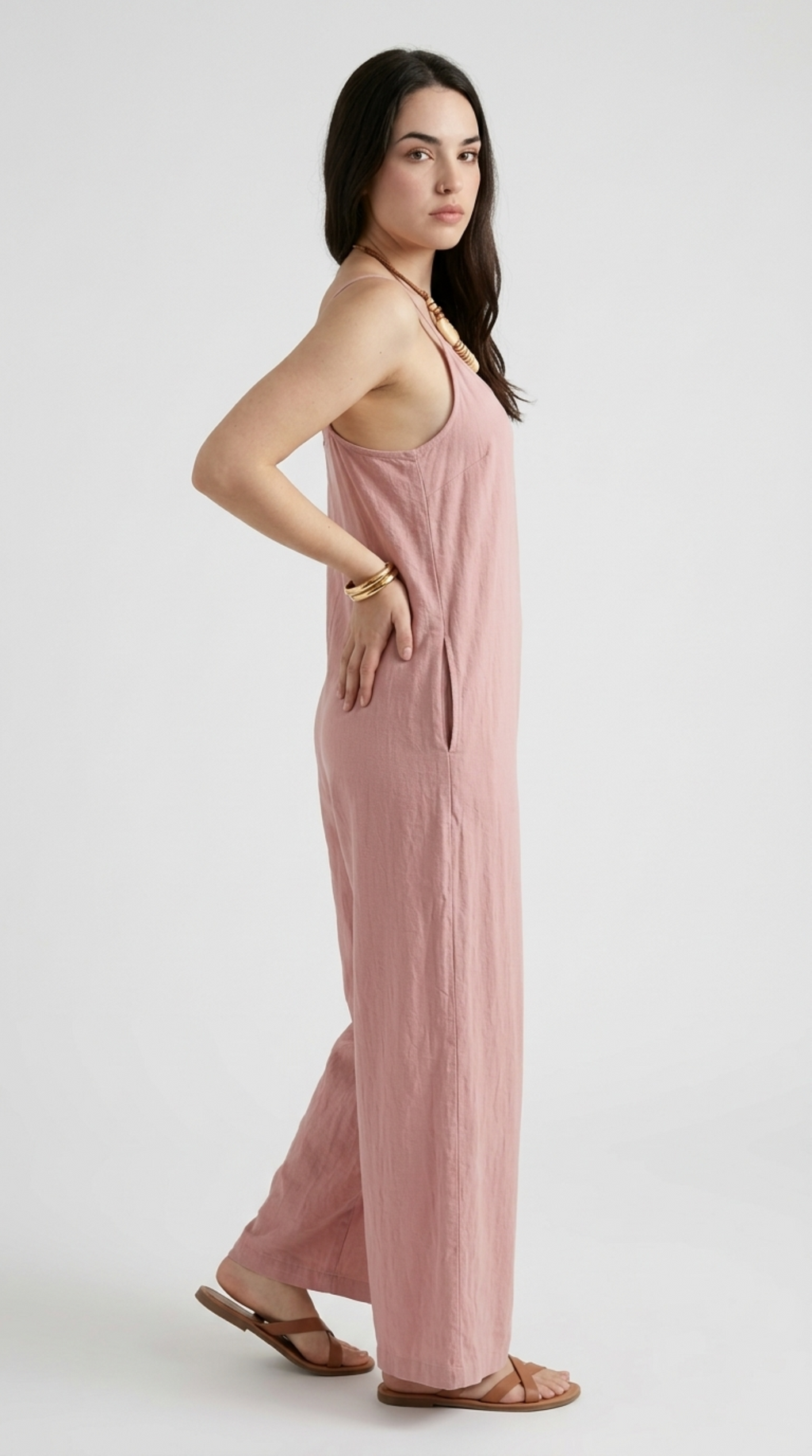 A woman modeling a dusty pink sleeveless jumpsuit with a relaxed, wide-leg silhouette and thin shoulder straps, paired with brown leather sandals and gold bracelets, against a plain light gray studio background, from Meleche Fashion or Meléche (short as (Mé)) and shoot taken by Meléche studio photography NYC-USA