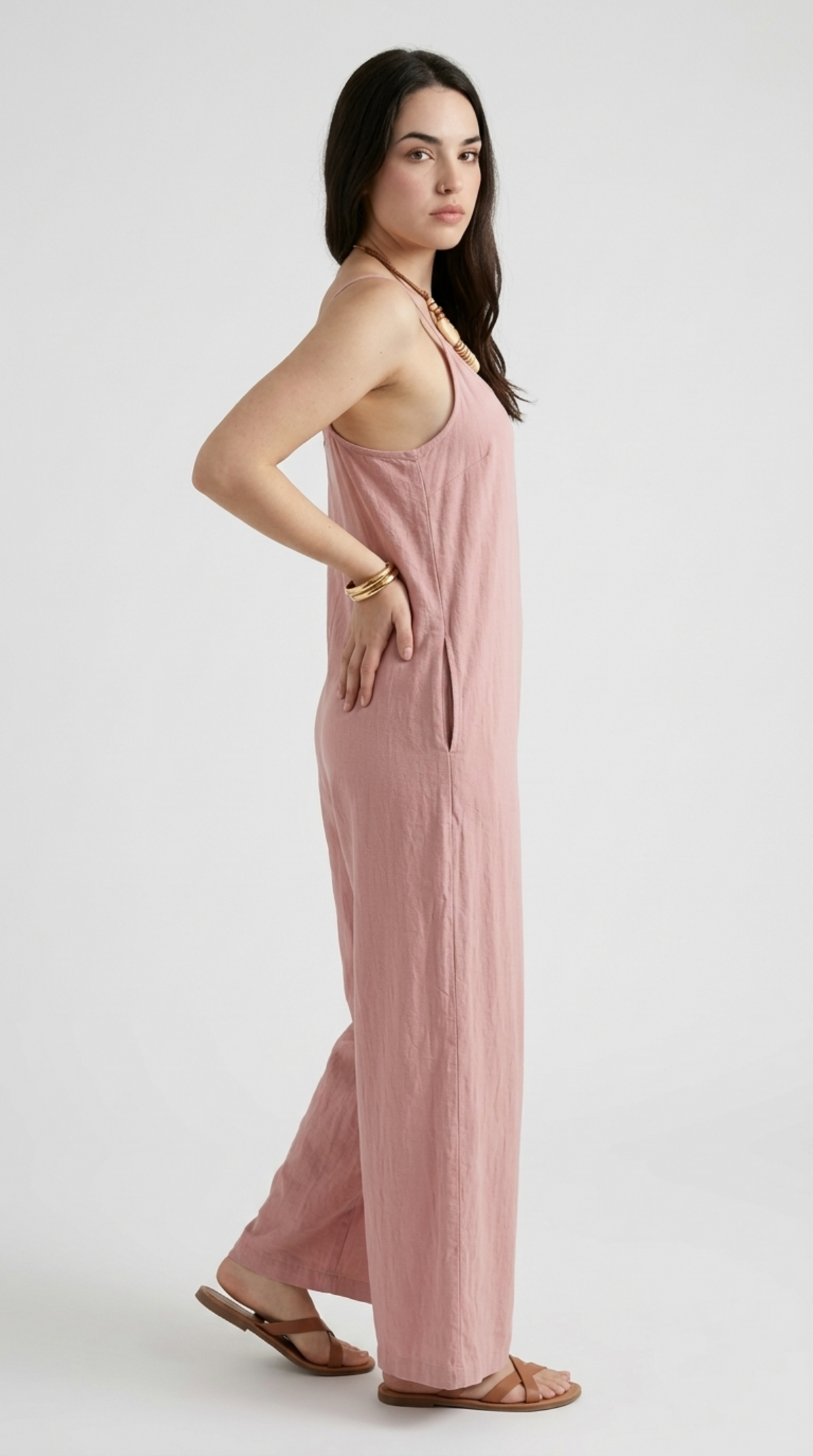 A woman modeling a dusty pink sleeveless jumpsuit with a relaxed, wide-leg silhouette and thin shoulder straps, paired with brown leather sandals and gold bracelets, against a plain light gray studio background, from Meleche Fashion or Meléche (short as (Mé)) and shoot taken by Meléche studio photography NYC-USA