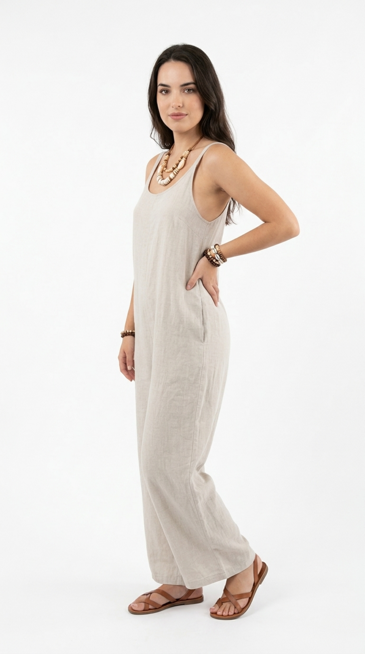 A woman modeling a light beige linen sleeveless maxi jumpsuit with wide legs, styled with a chunky gold necklace, bracelets, and brown leather sandals against a plain white studio background, from Meleche Fashion or Meléche (short as (Mé)) and shoot taken by Meléche studio photography NYC-USA