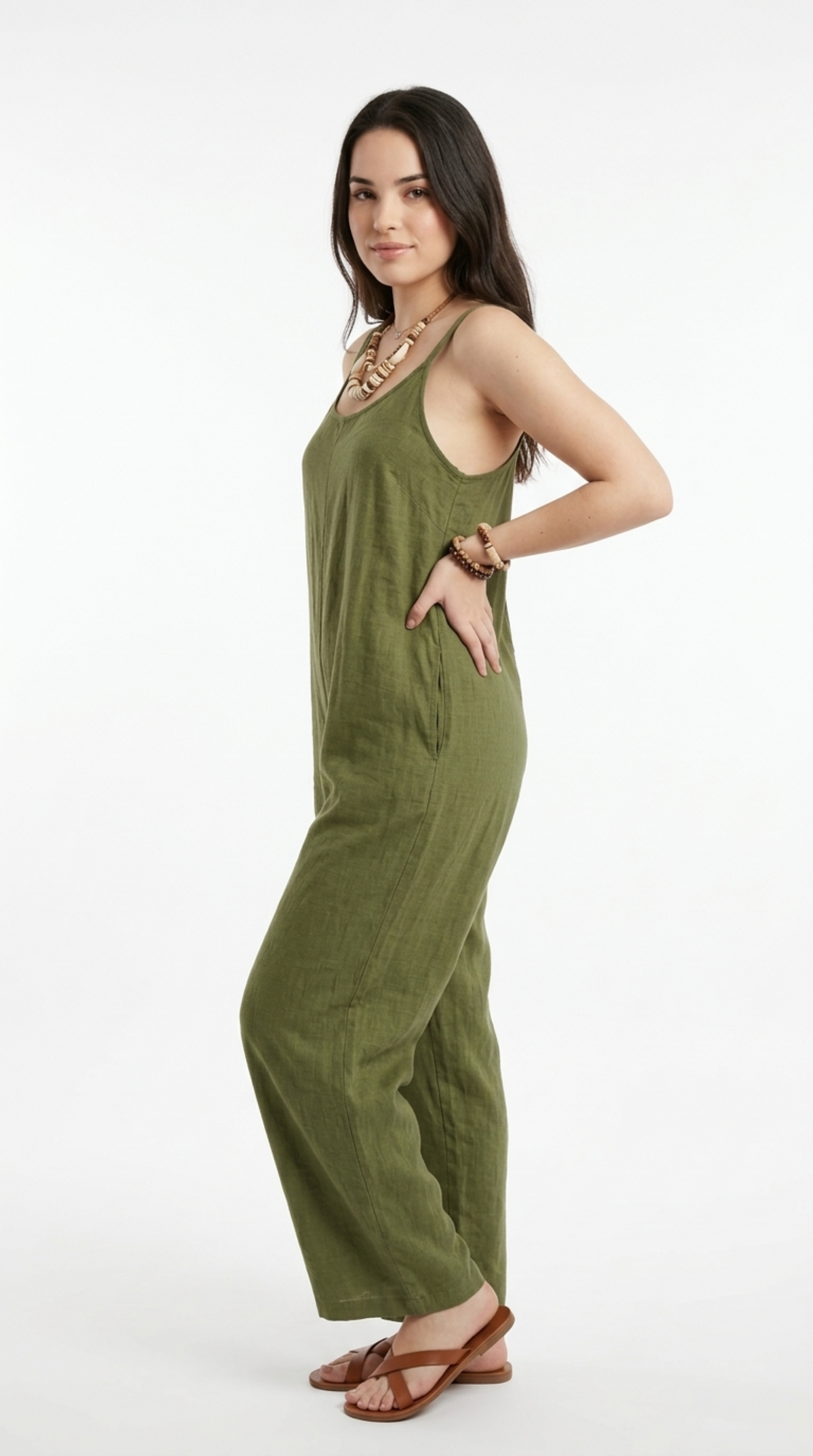 A woman modeling a sleeveless olive green linen jumpsuit with wide legs, styled with brown leather flat sandals and a chunky gold necklace, against a plain white studio background, from Meleche Fashion or Meléche (short as (Mé)) and shoot taken by Meléche studio photography NYC-USA