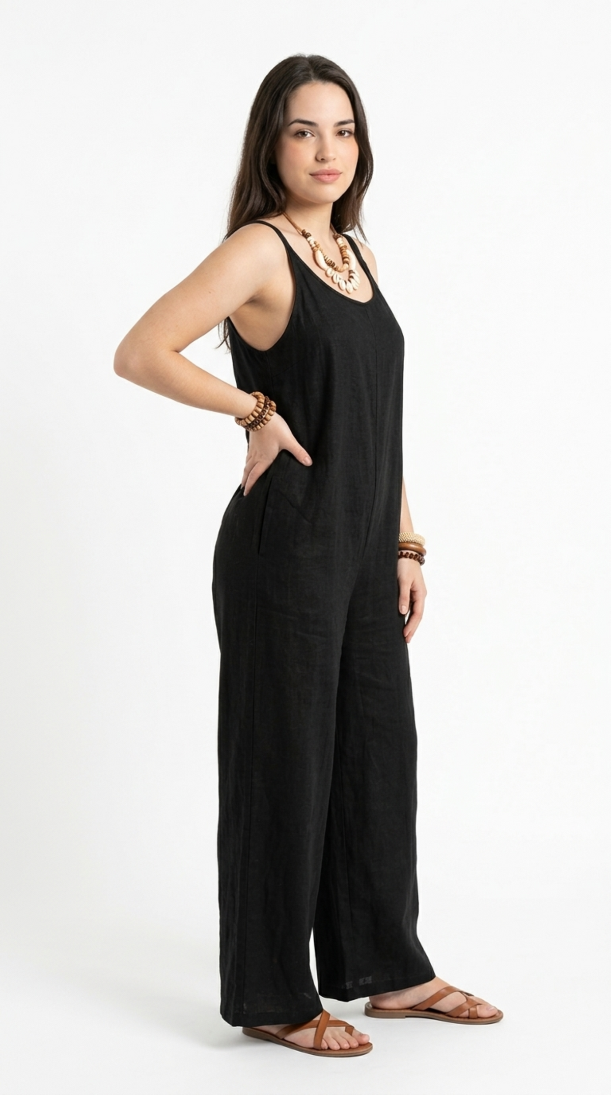 A woman modeling a black sleeveless wide-leg jumpsuit with side pockets, paired with brown leather sandals and a chunky statement necklace, photographed against a plain white studio background, from Meleche Fashion or Meléche (short as (Mé)) and shoot taken by Meléche studio photography NYC-USA