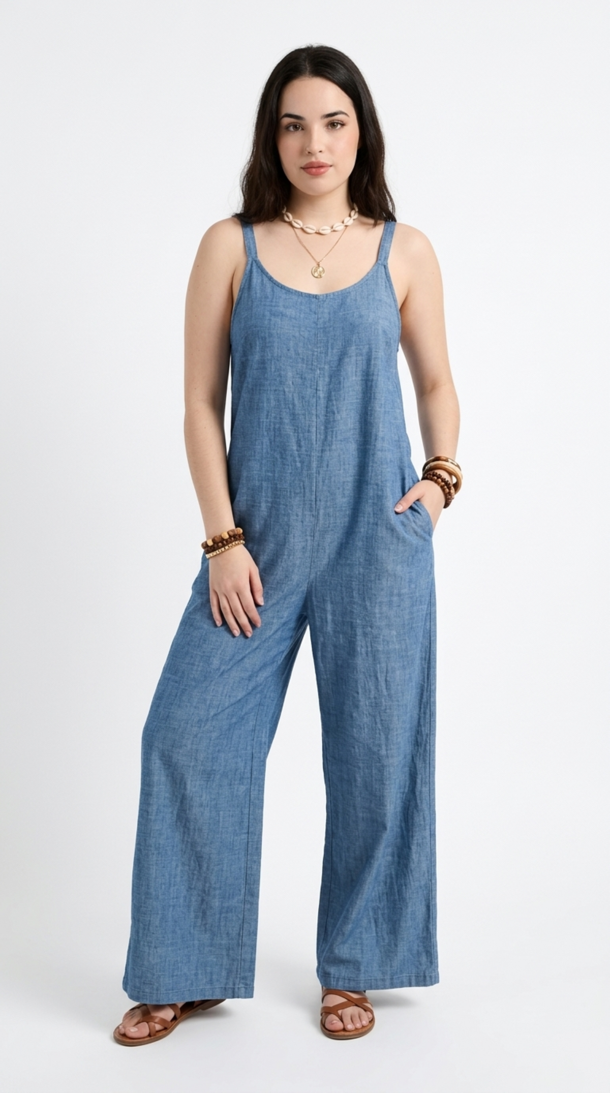 A woman modeling a blue wide-leg jumpsuit with thin straps and side pockets, paired with brown flat sandals and layered gold necklaces, against a plain white studio background, from Meleche Fashion or Meléche (short as (Mé)) and shoot taken by Meléche studio photography NYC-USA