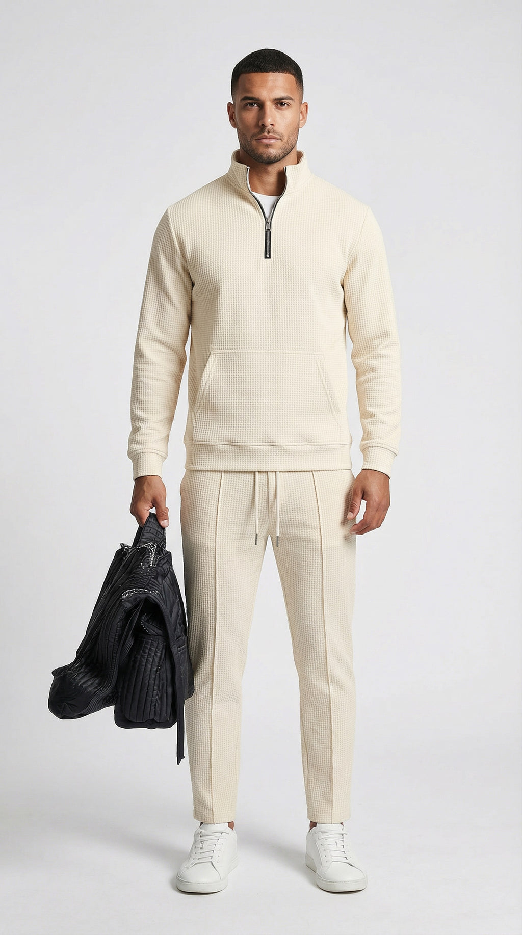Man wearing a beige tracksuit holding a black jacket on a white background
