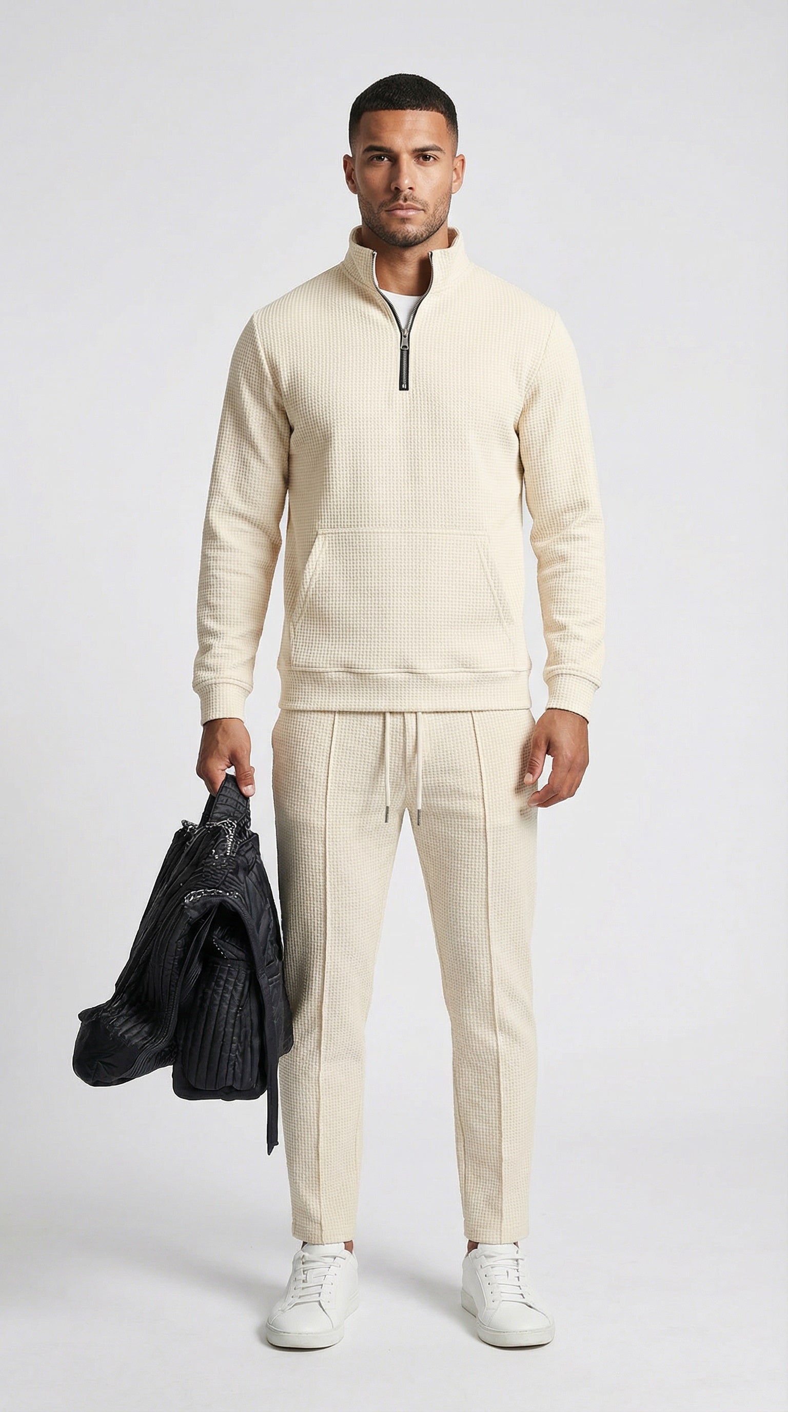 Man wearing a beige tracksuit holding a black jacket on a white background