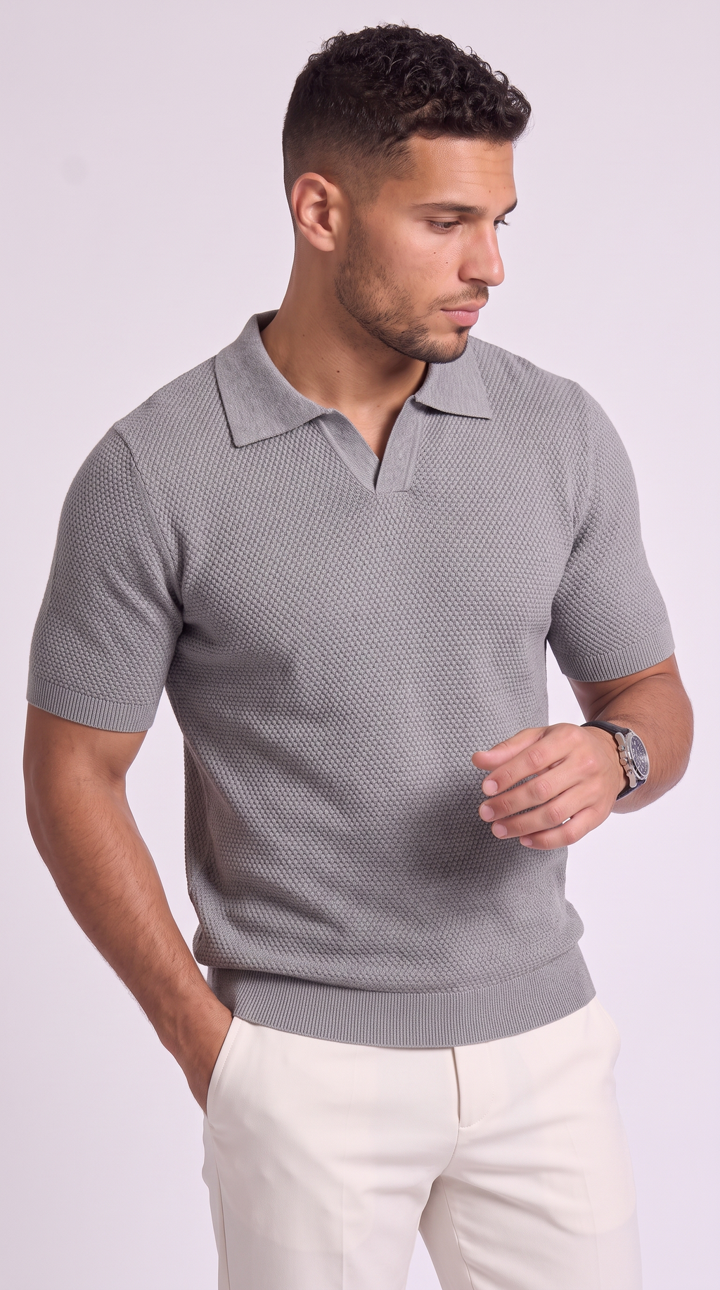 A man wearing a gray short-sleeve polo shirt with a textured knit pattern and white trousers, standing against a light gray studio background, from Meleche Fashion USA or Meléche (short as (Mé))