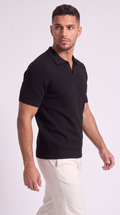 A man wearing a black short-sleeve polo shirt with a textured knit pattern and white trousers, photographed against a light gray studio background, from Meleche Fashion USA or Meléche (short as (Mé))