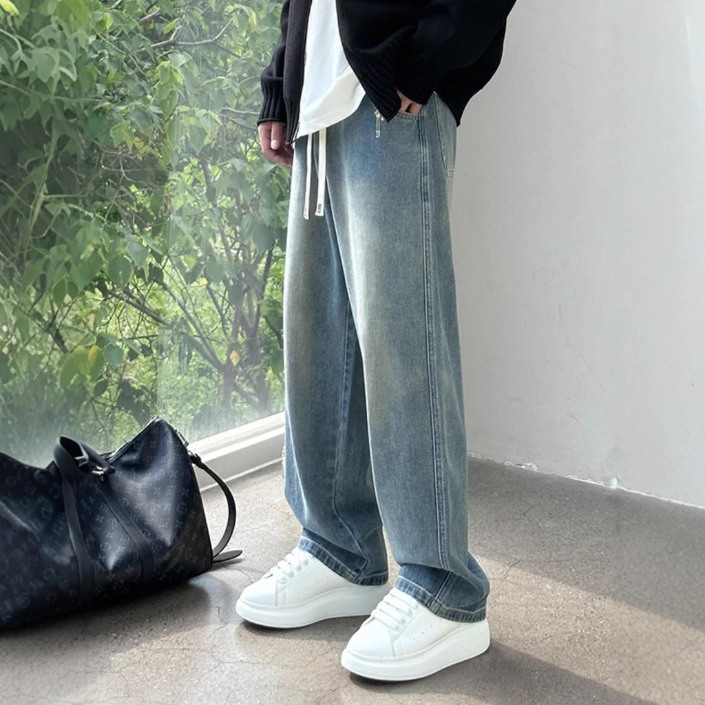 Person wearing blue jeans and white sneakers standing next to a black bag indoors.