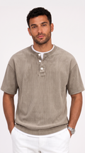 A man wearing a taupe ribbed short-sleeve henley shirt with white buttons and a white undershirt, paired with white pants, against a light gray studio background, from Meleche Fashion USA or Meléche (short as (Mé))