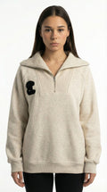 Sophie Rain wearing a beige pullover with a black patch from Meleche or Meléche (short as (Mé)) on a white background