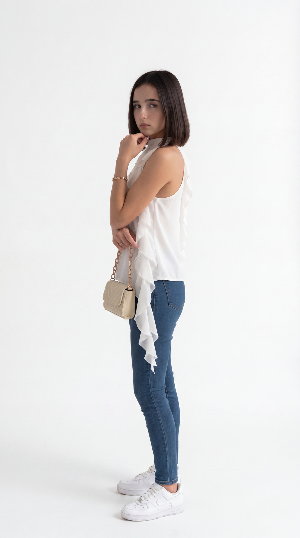 A woman posing in a white sleeveless ruffled top, blue skinny jeans, and white sneakers, holding a small beige handbag against a plain white studio background, from Meleche Fashion USA or Meléche (short as (Mé))