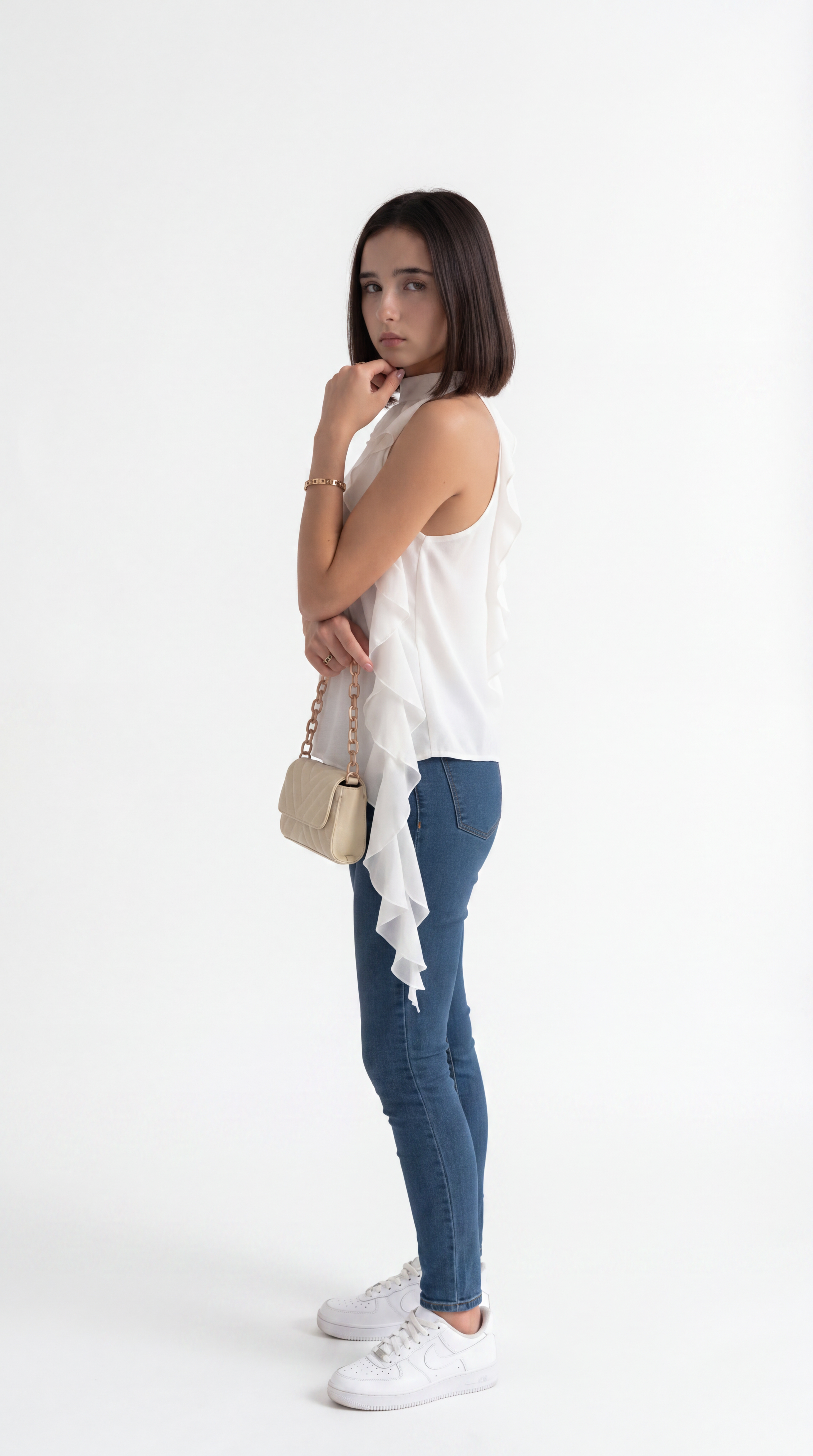 A woman posing in a white sleeveless ruffled top, blue skinny jeans, and white sneakers, holding a small beige handbag against a plain white studio background, from Meleche Fashion USA or Meléche (short as (Mé))