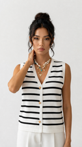 A woman modeling a sleeveless knit vest with horizontal black and white stripes, featuring a V-neckline and three gold buttons, paired with white trousers and layered gold necklaces, against a plain light gray background.